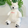 A white plush polar bear stuffed animal with black nose and paw pads, sitting in front of green tropical leaves.