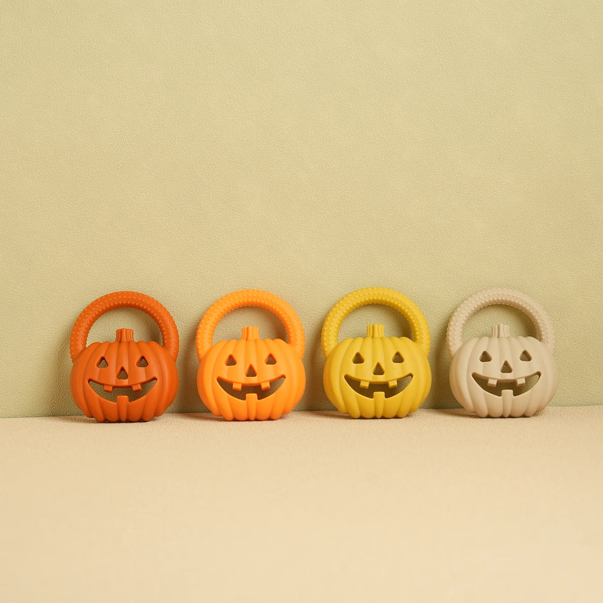 A row of four silicone pumpkin baby teethers in orange, yellow, and white are displayed on a light background.