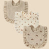 A set of three light beige cotton muslin baby bibs with ruffled edges and decorative patterns