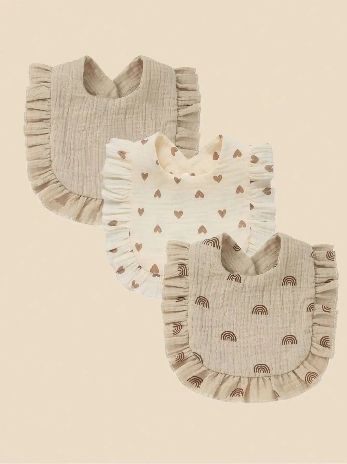 A set of three light beige cotton muslin baby bibs with ruffled edges and decorative patterns