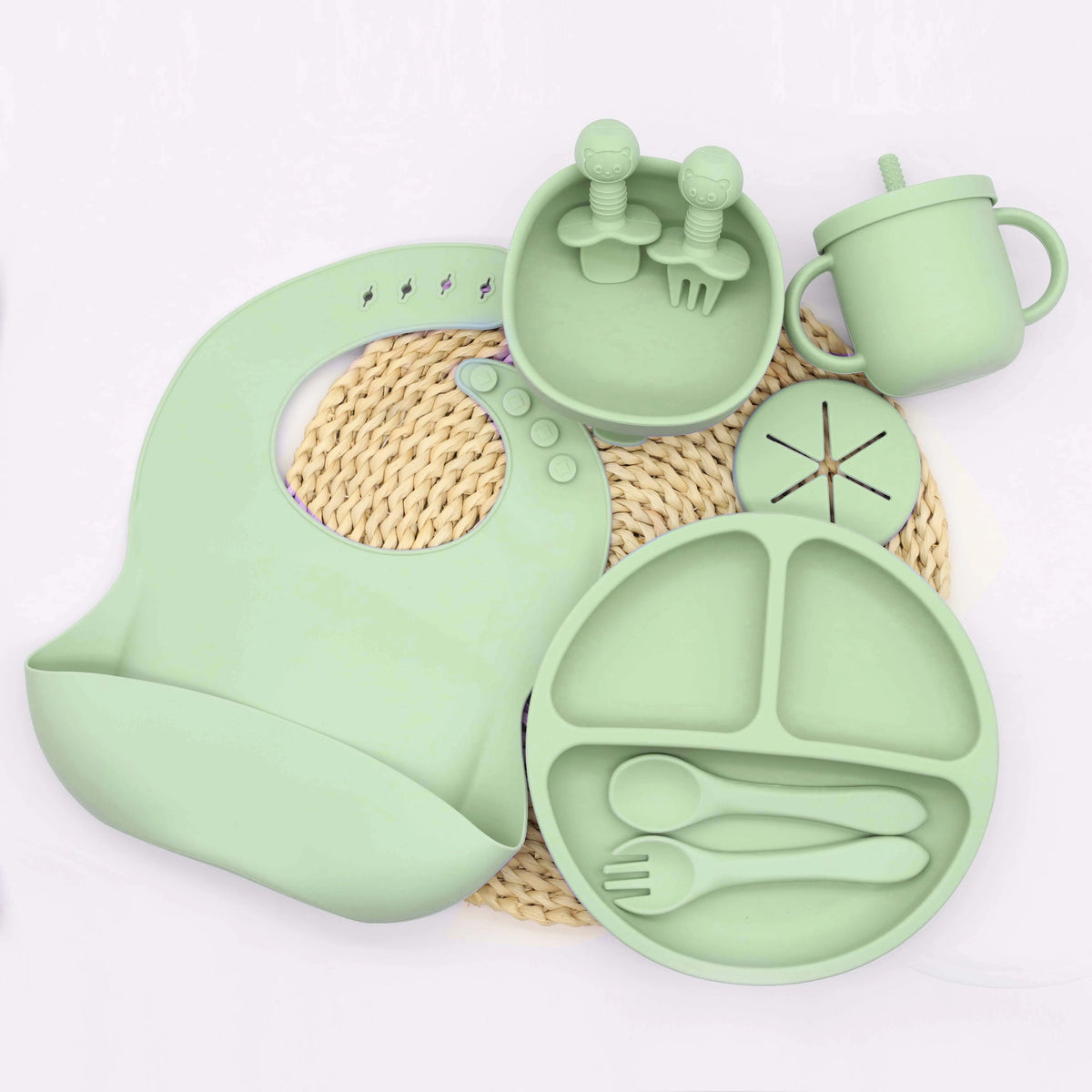 A light green silicone tableware set for babies, including cups, plates, and utensils, resting on a woven mat.