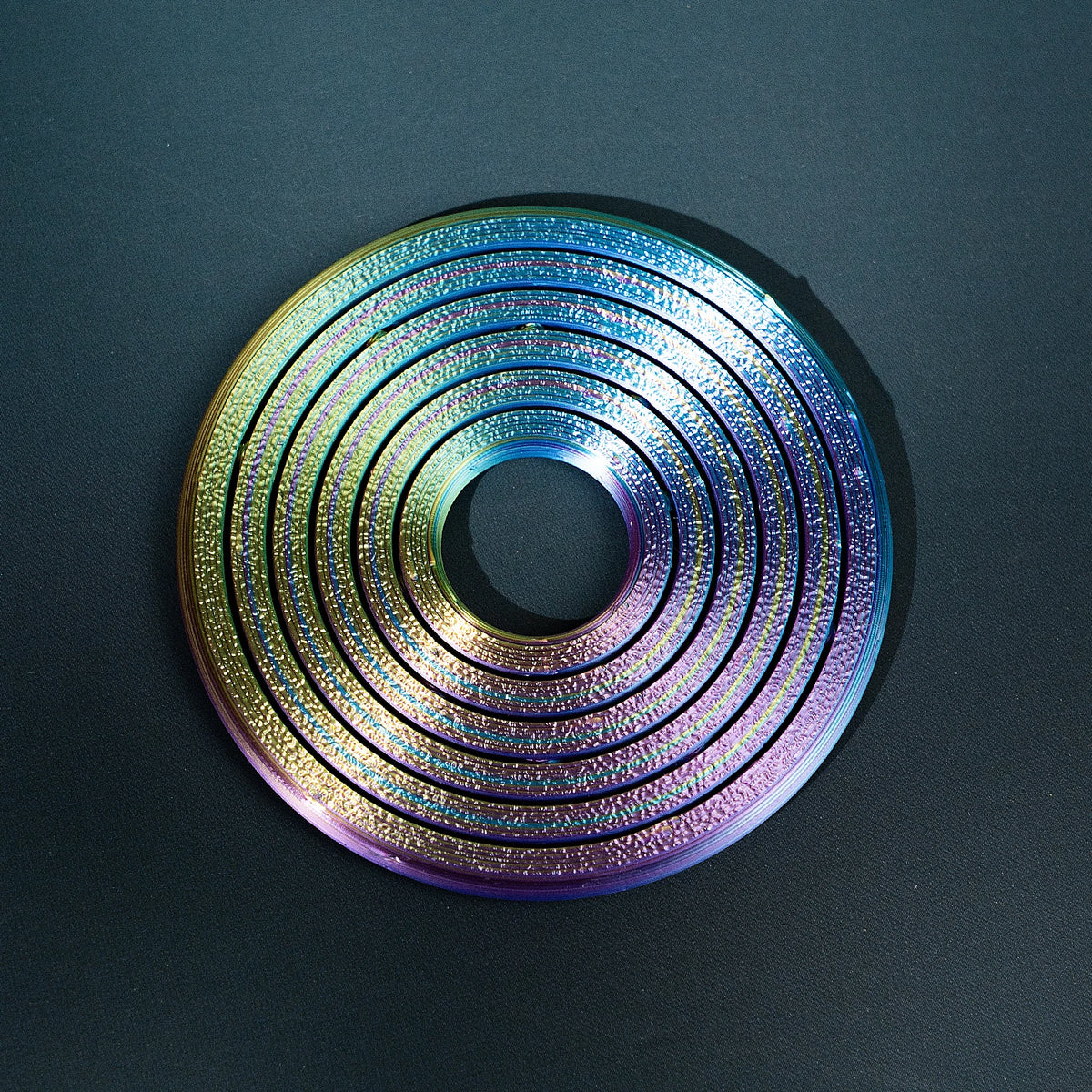 A close-up of a 3D printed rainbow spinner ring with concentric grooves and iridescent colors on a dark surface.