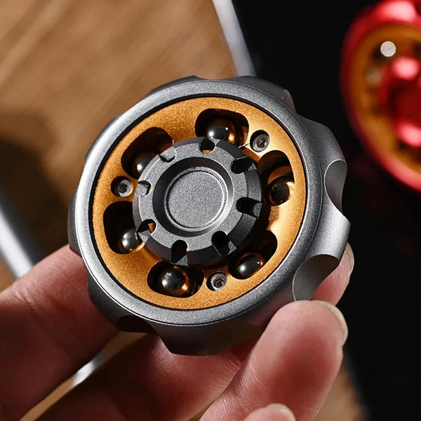 A hand holds a metal fidget toy with a silver outer shell and gold inner ring featuring multiple small, dark-colored componen