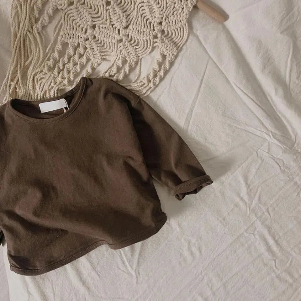 A brown long sleeve cotton tee is laid on a white surface next to a cream-colored knitted garment.