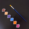 A set of twelve colored acrylic paint pots arranged in a line with a paintbrush next to them.