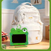 A white backpack with a green frog-shaped LCD drawing tablet keychain resting on a wooden surface.
