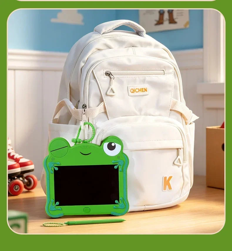 A white backpack with a green frog-shaped LCD drawing tablet keychain resting on a wooden surface.