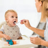 A woman is helping a baby put food on a silicone spoon while playing with colorful wooden blocks.