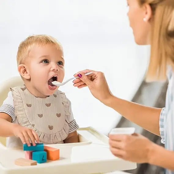 A woman is helping a baby put food on a silicone spoon while playing with colorful wooden blocks.