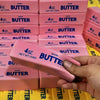 A hand holds a pink butter stick fidget toy packaged in boxes labeled Butter
