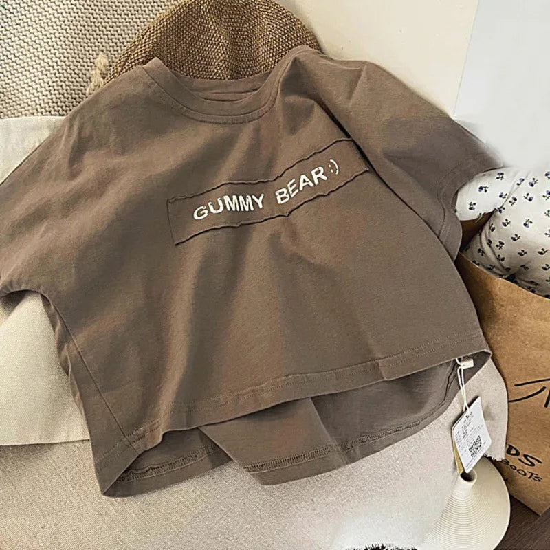 A plain taupe short-sleeve t-shirt with the text GUMMY BEAR printed on the chest, displayed on a light surface.