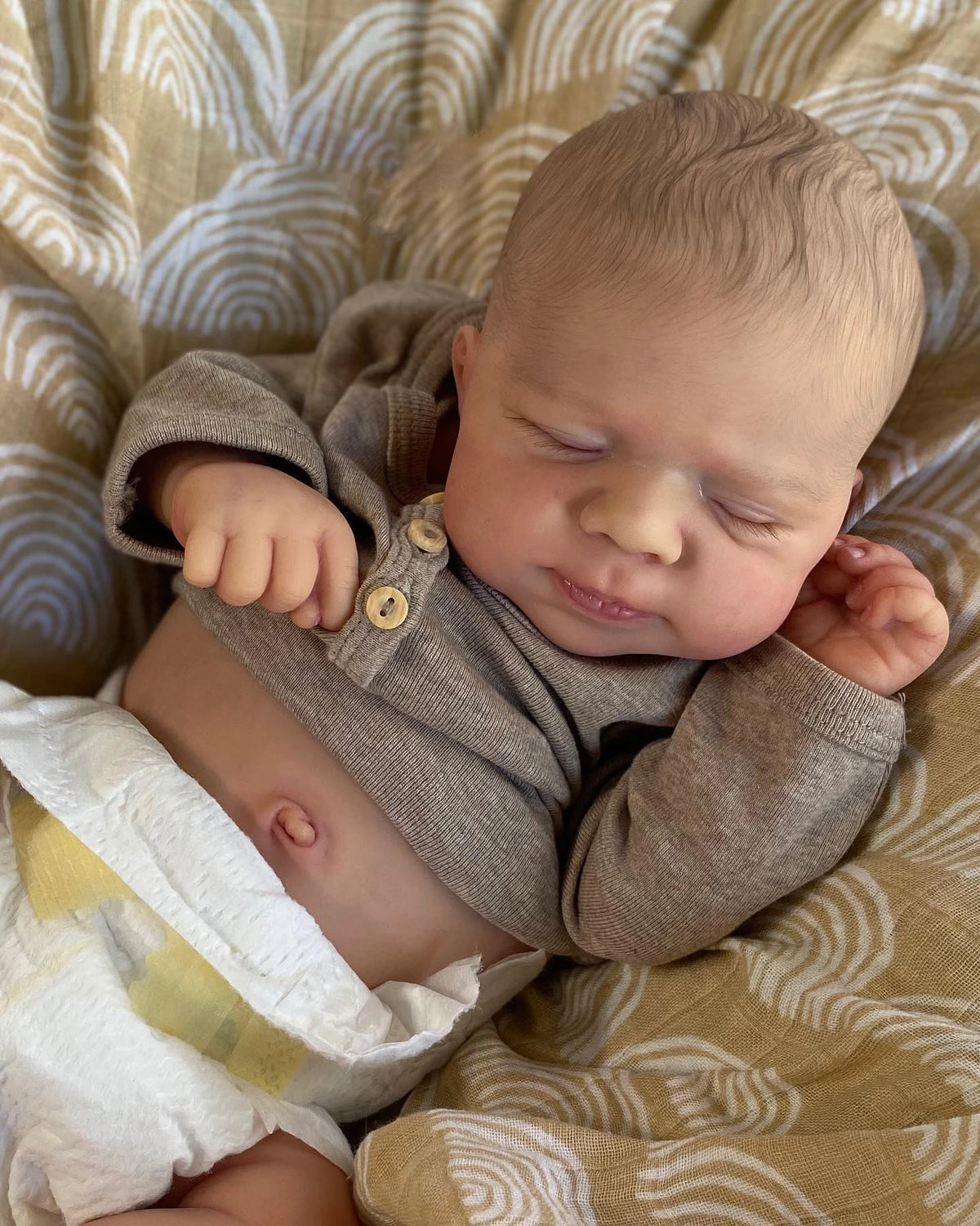 A newborn silicone reborn doll is lying on a patterned fabric, sleeping with eyes closed.