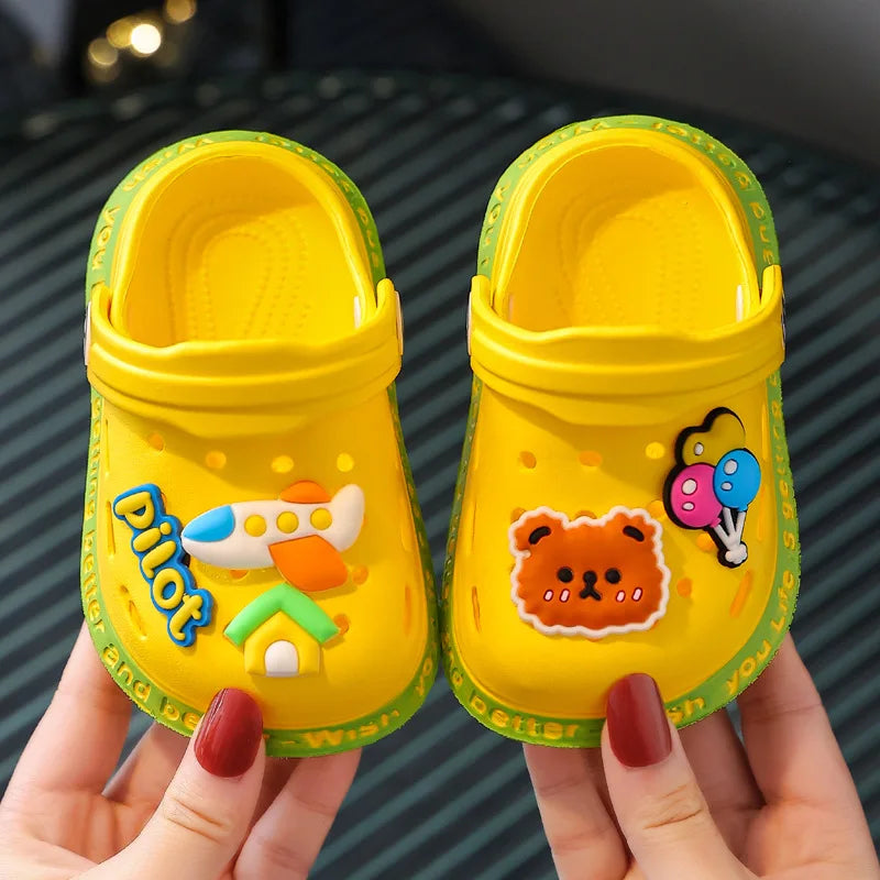 A pair of bright yellow lightweight children's clogs with orange and green decorations being held by a hand.