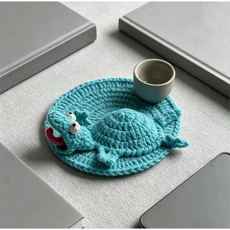 Handmade crochet lizard coaster in teal color resting on a light gray surface with a small ceramic cup.