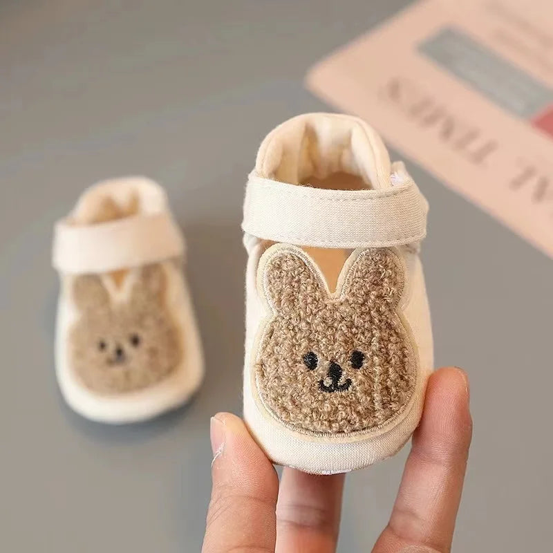 A pair of adorable bear soft sole first walker shoes for babies in cream and brown colors being held by a hand.