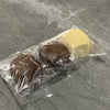 Un sac plastique transparent contient deux pains décoratifs en tissu brun et un jaune sur une surface grise.