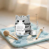 A handmade crochet cat figurine with gray and white stripes sitting on a light blue towel next to a wooden spoon.