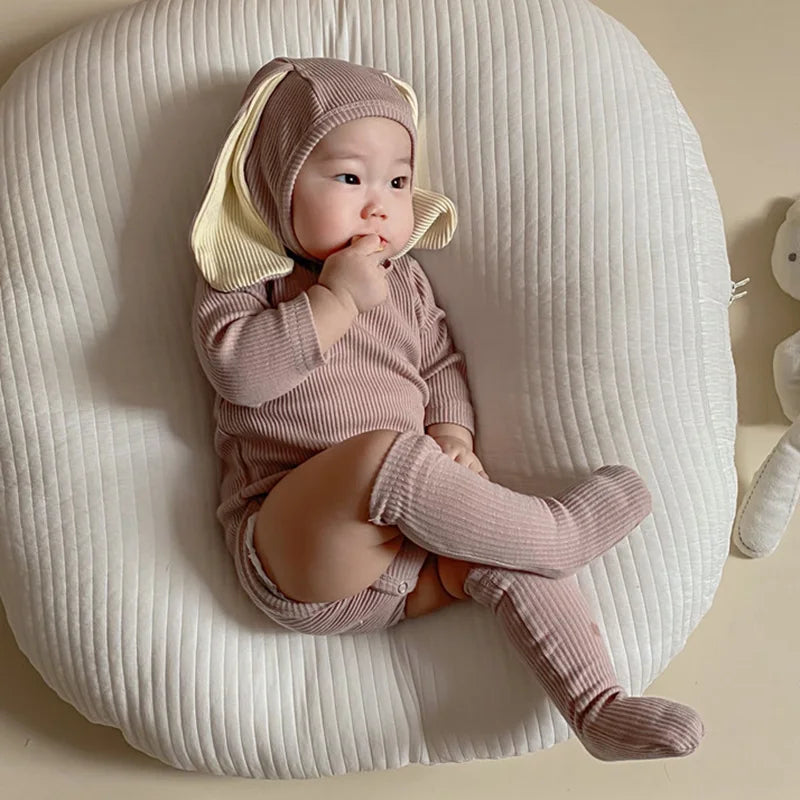 A baby wearing a pink rabbit romper set with a matching hat and socks, lying on a white textured surface.