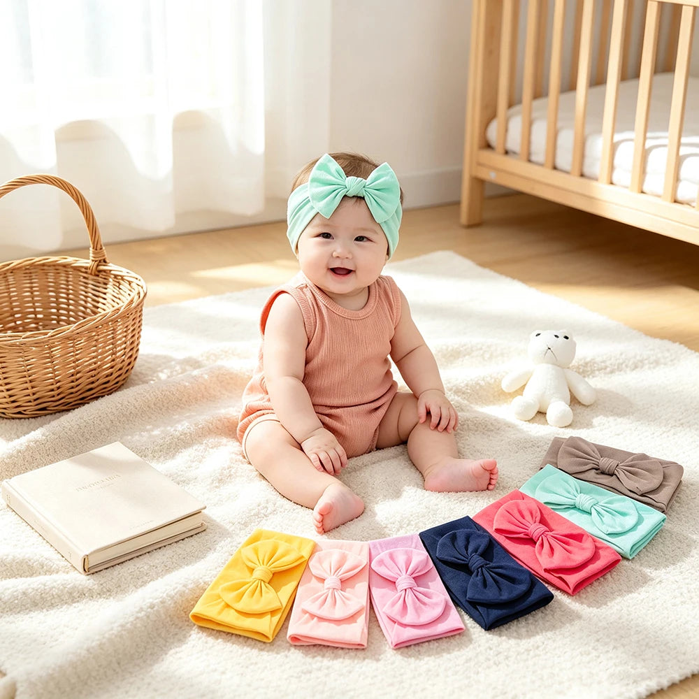 A baby girl wearing a pink bow headband sitting on a white rug surrounded by various colored fabric bows.