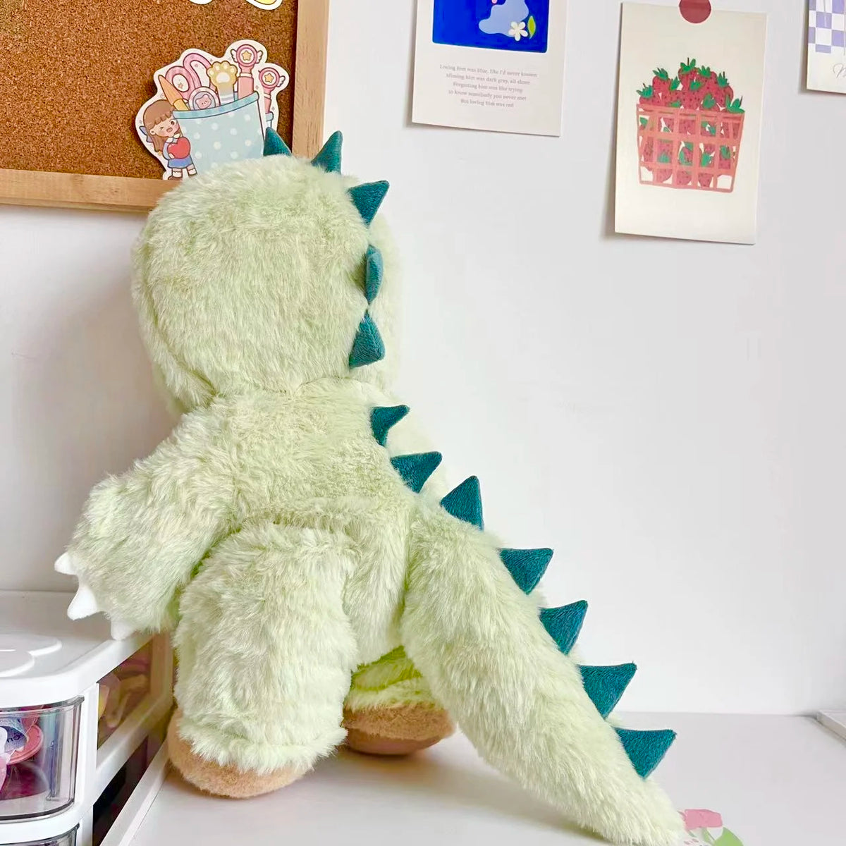 A soft, light green dinosaur plush toy with spiky dorsal fins is displayed against a white wall.