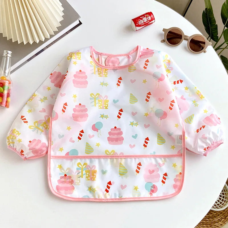 A full-sleeve waterproof baby bib with a pink and yellow cartoon design featuring stacked cakes, stars, and hearts.