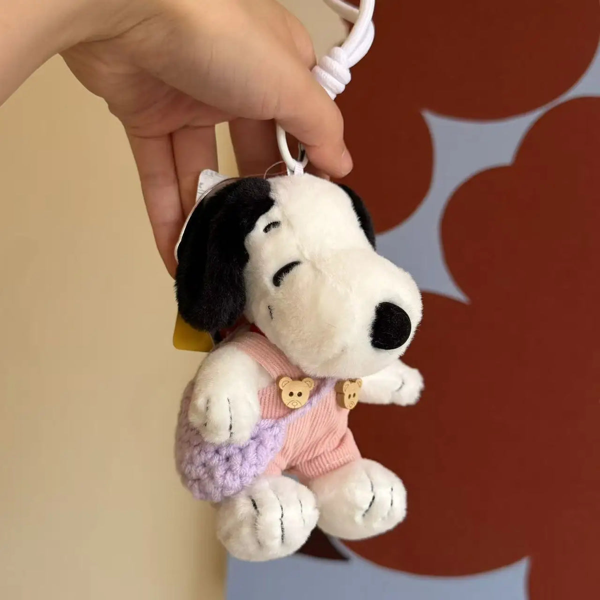A white and black Snoopy plush keychain charm is being held by a hand against a brown patterned background.