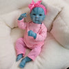 A handcrafted reborn baby doll with a blue silicone vinyl body and pink hat sitting on a white fluffy surface.