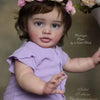 A close-up photo of a baby doll with dark hair wearing a purple dress and a pink floral headband.