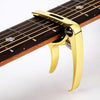 A gold-colored metal guitar capo with a pick holder is shown clamped onto a wooden guitar fretboard.