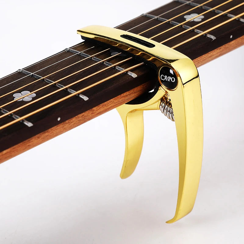A gold-colored metal guitar capo with a pick holder is shown clamped onto a wooden guitar fretboard.