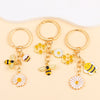 Three gold enamel flower keychains featuring bees and daisies on a light beige background.