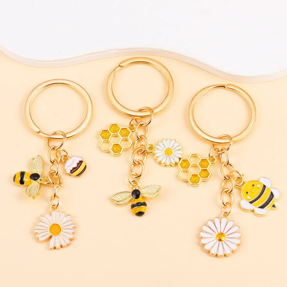 Three gold enamel flower keychains featuring bees and daisies on a light beige background.