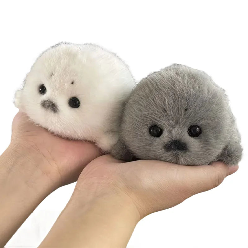 A pair of adorable grey seal plush keychains, one white and one grey, held in a person's hand.