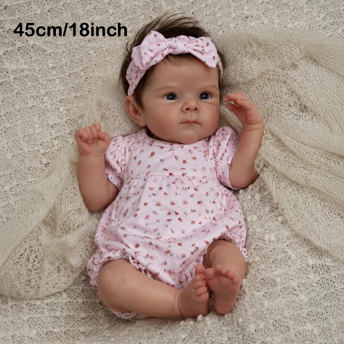 A newborn baby doll with hand-rooted hair wearing a pink floral outfit and a matching bow, lying on a white textured fabric.