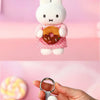 A white Miffy plush keychain charm with a pink dress and an orange donut, being held by a hand.