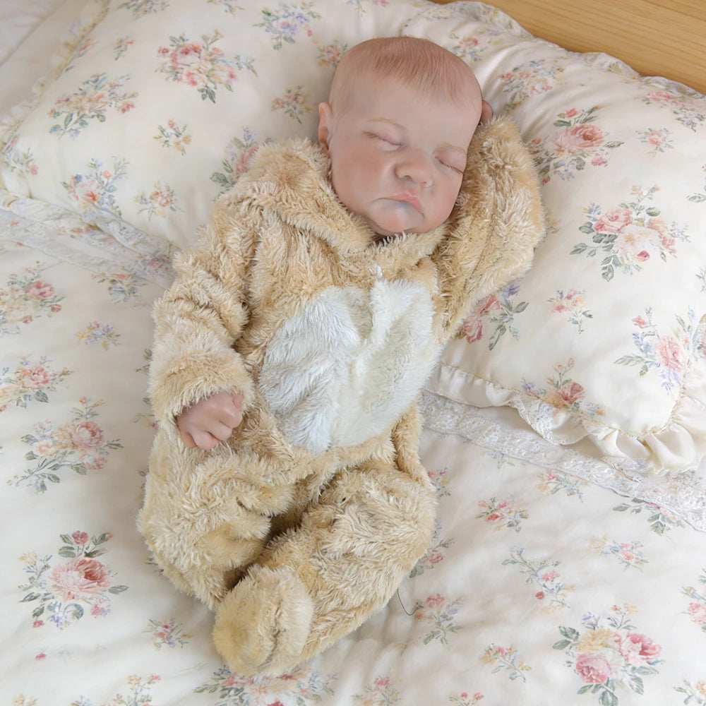 A realistic 18-inch vinyl reborn baby doll wearing a fluffy bear outfit sleeping on a floral patterned fabric.