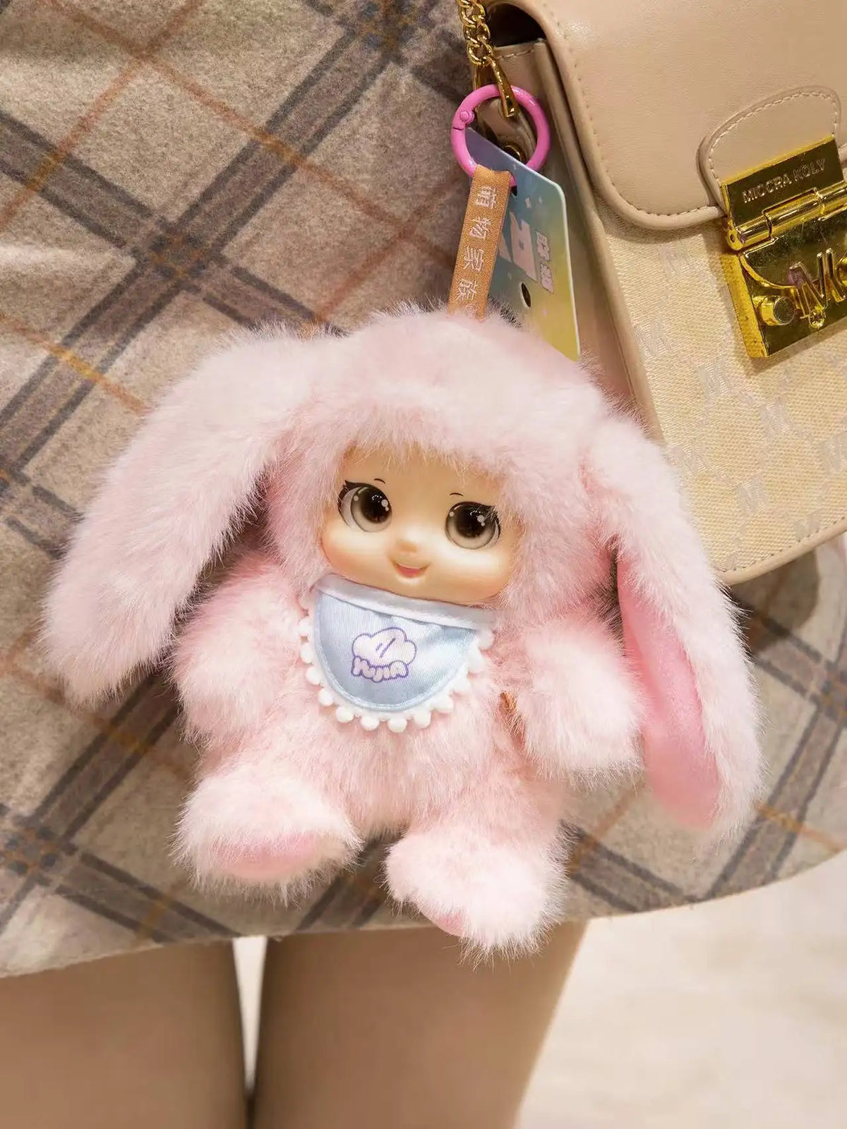 A pink fluffy rabbit plush pendant hanging from a gold chain on a beige bag.