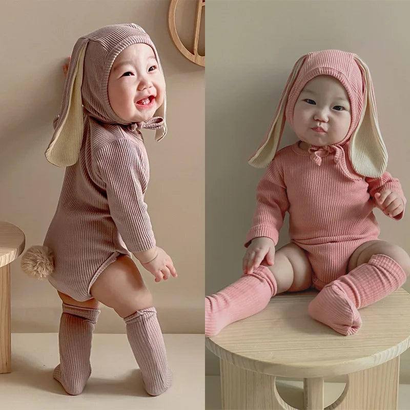 A baby wearing a pink rabbit romper set with a matching hat and socks sitting on a wooden surface.