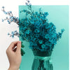 A hand holds a bouquet of blue flowers in a glass vase against a teal background.