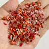 A hand holds a pile of natural tumbled stone chips in various shades of pink, red, and amber for jewelry making or decor.