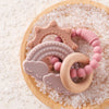 A set of food-grade silicone planet teethers in pink and beige are displayed on a wooden board surrounded by clear, sparkling