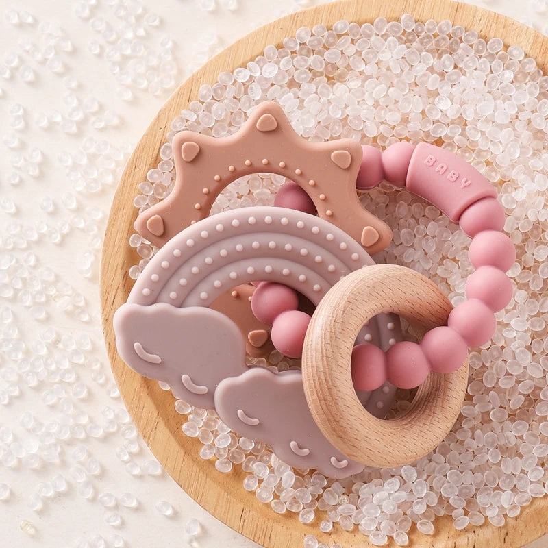A set of food-grade silicone planet teethers in pink and beige are displayed on a wooden board surrounded by clear, sparkling