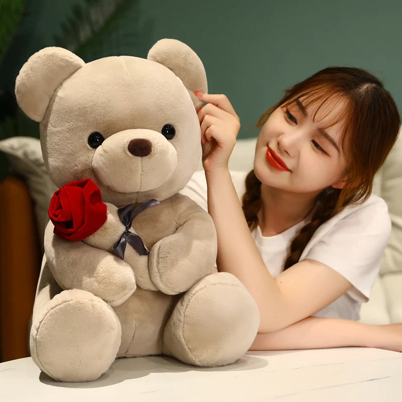 A young woman is holding a light brown teddy bear plush toy with a red rose in a soft indoor setting.