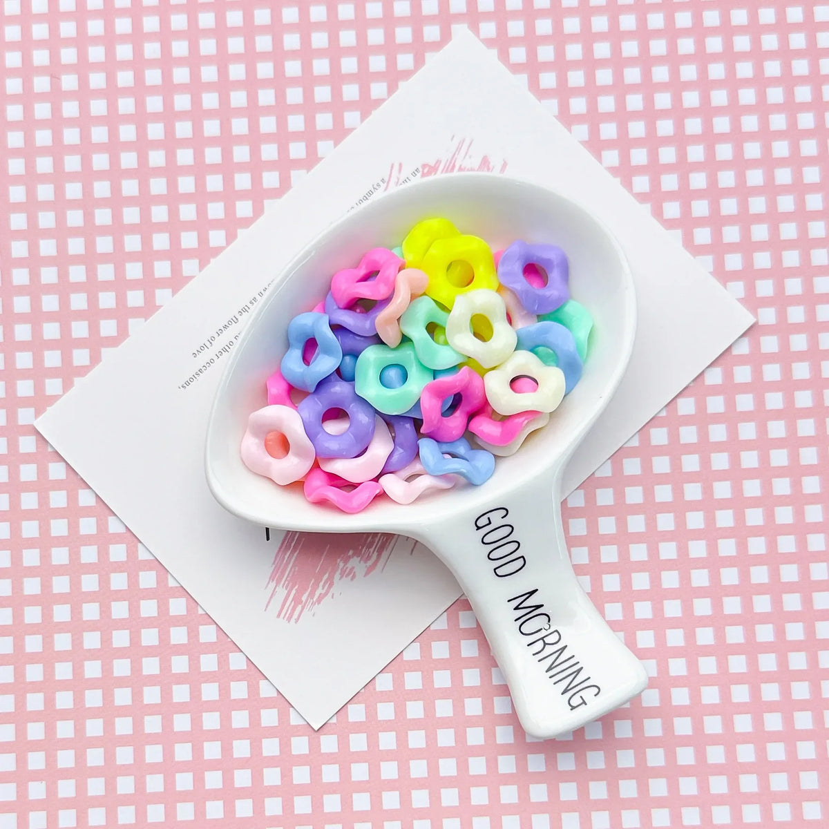 A white spoon holds colorful pastel wavy acrylic beads in various shades of pink, purple, and yellow on a pink checkered back