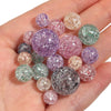 A hand holds a collection of round, loose glitter cracked acrylic beads in various shades of purple, pink, and green.