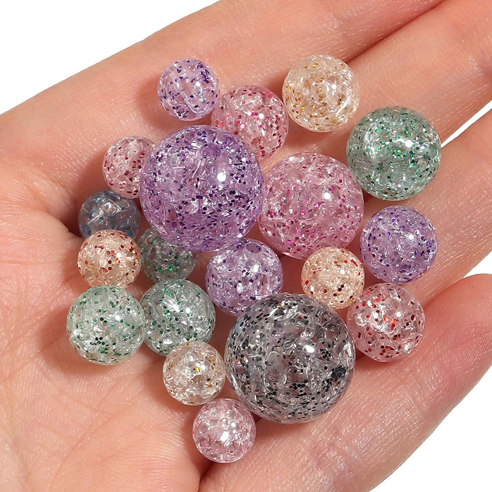 A hand holds a collection of round, loose glitter cracked acrylic beads in various shades of purple, pink, and green.