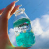 A hand holds a clear bottle filled with bright blue liquid, labeled Chiboki, against a cloudy blue sky.