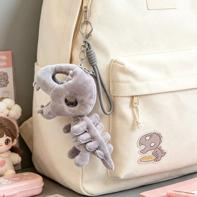 A light beige backpack features a gray dinosaur skeleton charm with a removable hood attached to the zipper pull.