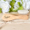 A light wooden baby brush and comb set with the name Sophie engraved on the handle, resting on a beige cloth.