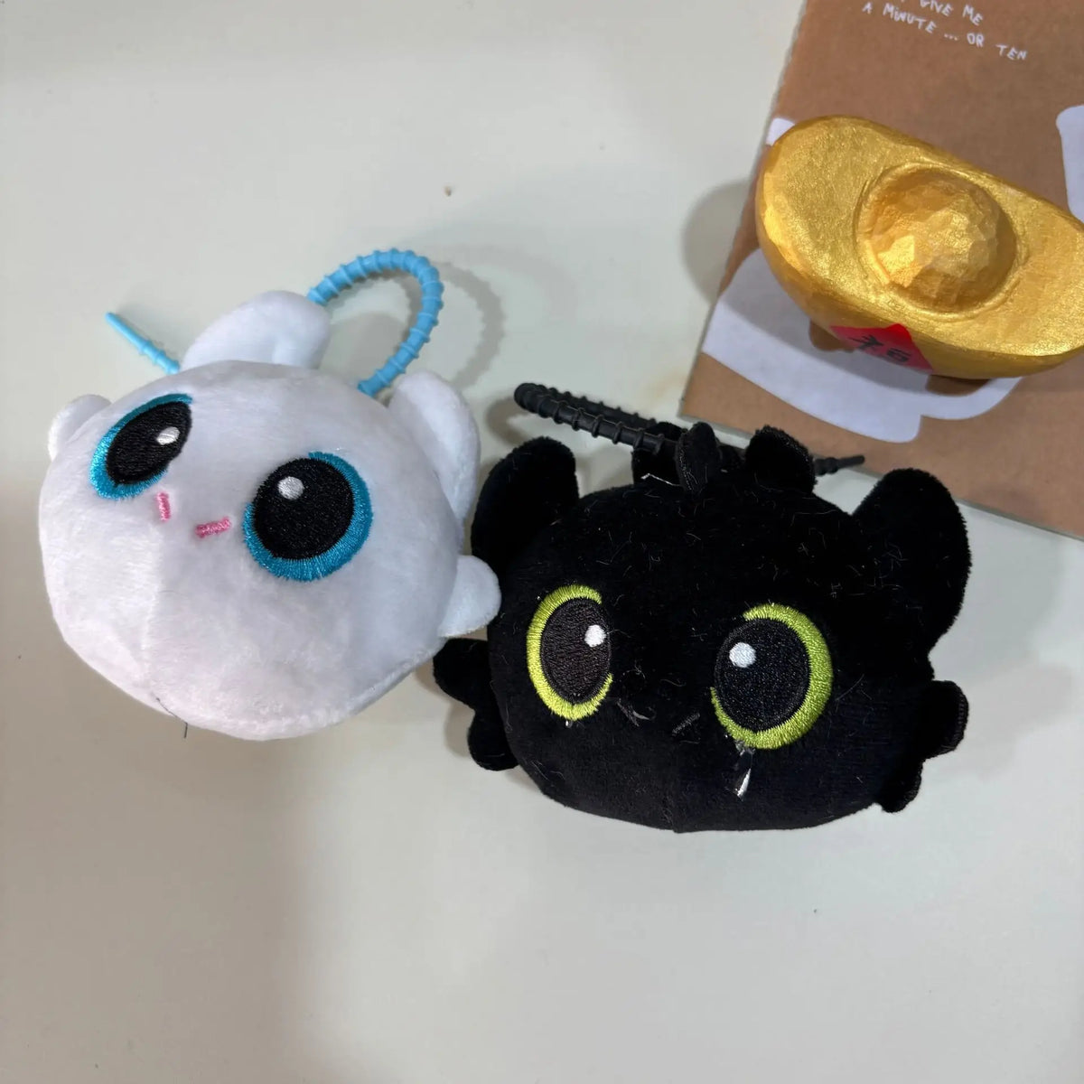 A white plush keychain of Toothless with blue accents is shown next to a black plush version.